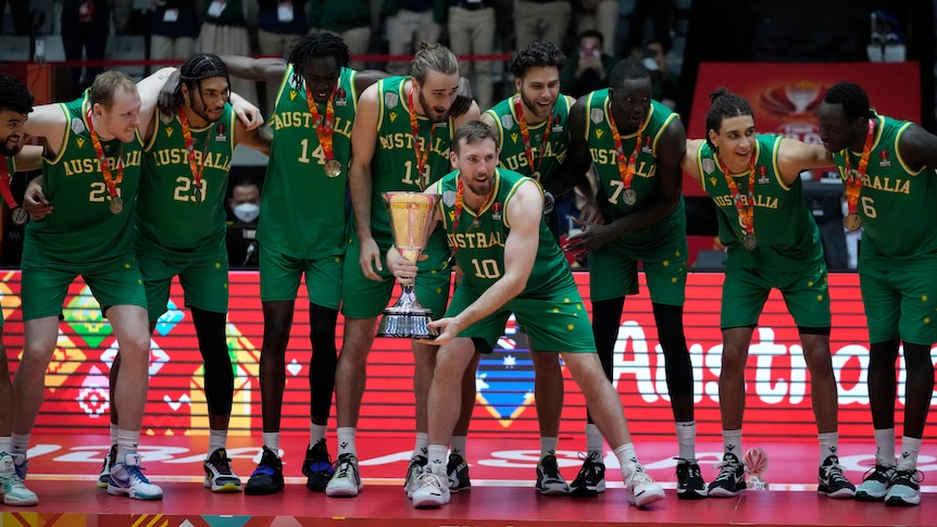 Australia's men's basketball team the Boomers lean in together on court as one player holds a trophy after a tournament.