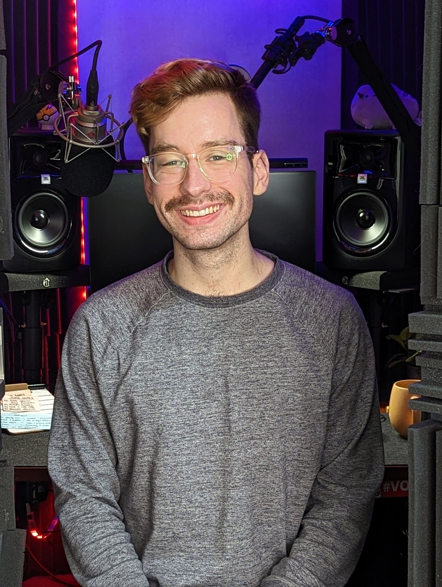 A moustached man with glasses stands in a sound studio.