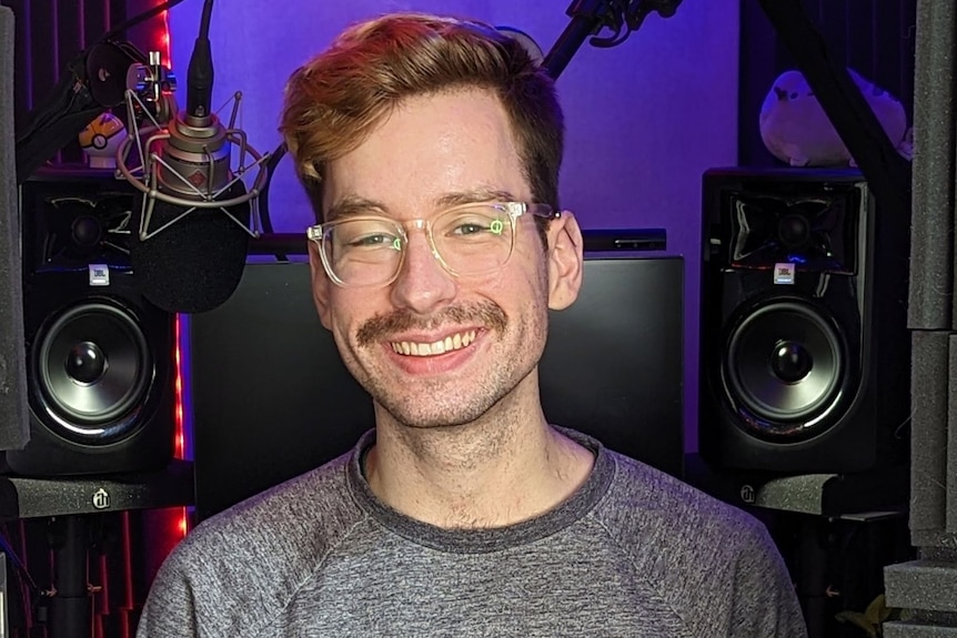 A moustached man with glasses stands in a sound studio.