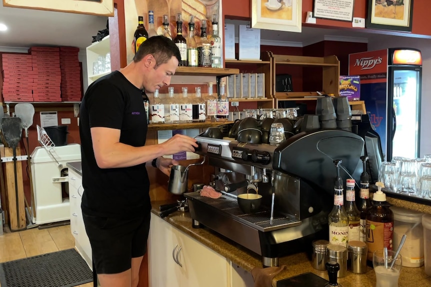 A man in black shirt and shorts stands in front of a coffee machine frothing some milk