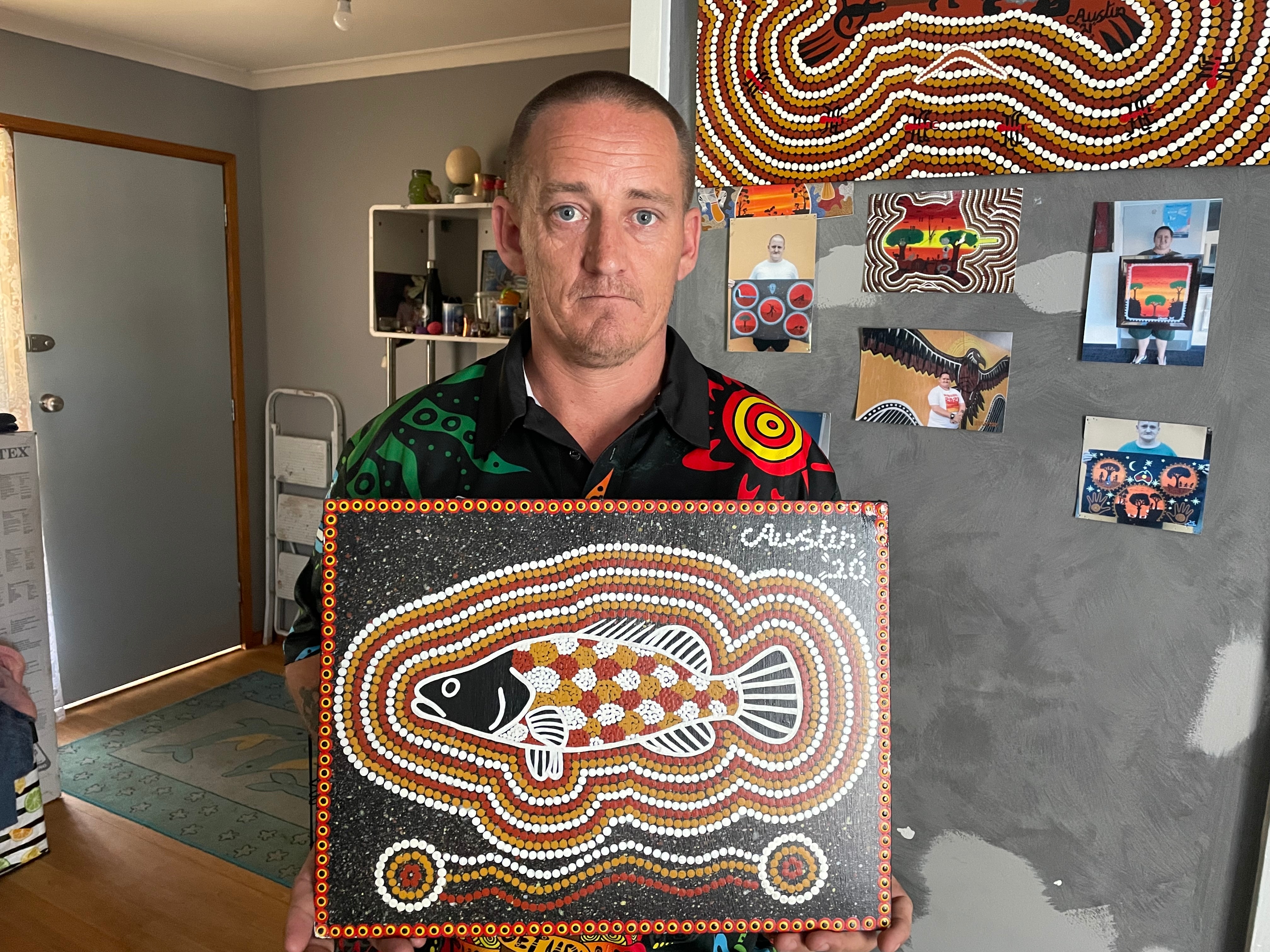 A man looks at the camera with a straight face. He holds an Aboriginal dot painting of a fish.