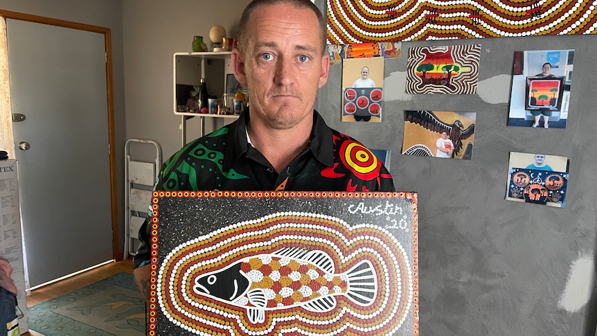 A man looks at the camera with a straight face. He holds an Aboriginal dot painting of a fish.