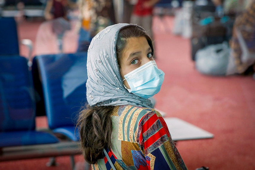 A young girl in a blue face mask and headscarf looking over her shoulder, making eye contact with the camera
