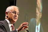 Malcolm Turnbull gestures as he speaks at a podium with a televised image of him on a large screen behind.