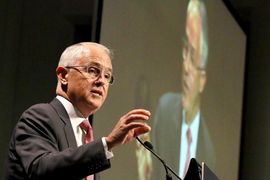 Malcolm Turnbull gestures as he speaks at a podium with a televised image of him on a large screen behind.