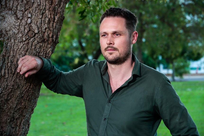 A man looks into the distance and leans against a tree.