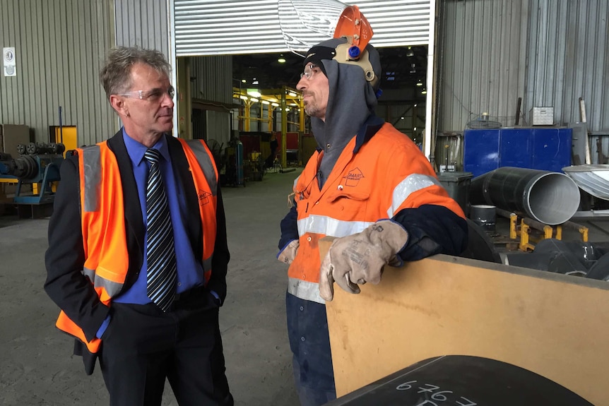 A man wearing an orange vest speaks to another man with a welder's helmet on