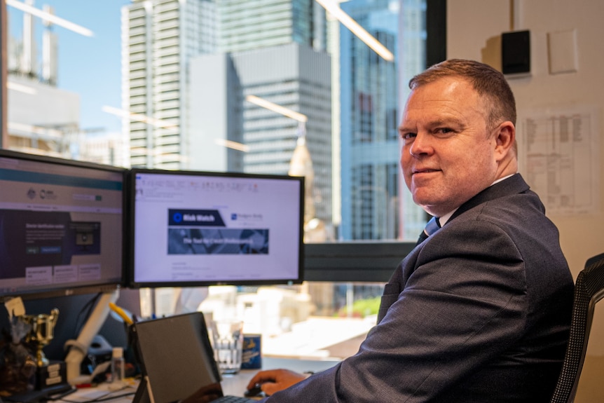 a man smiles at a camera in front of a computer with the words risk watch on the screen