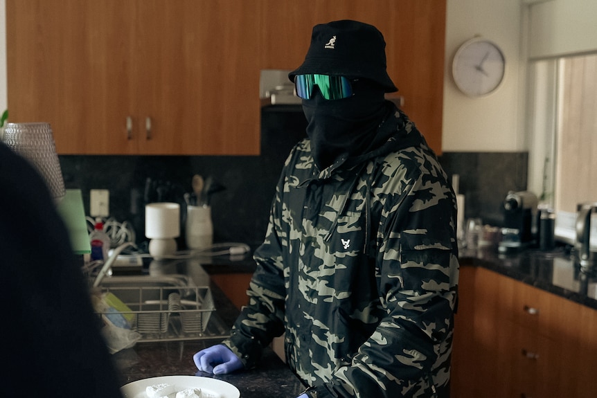 A man covered head to toe, wearing a hat, sunglasses, ski mask and gloves, stands in a kitchen with cocaine on a plate.