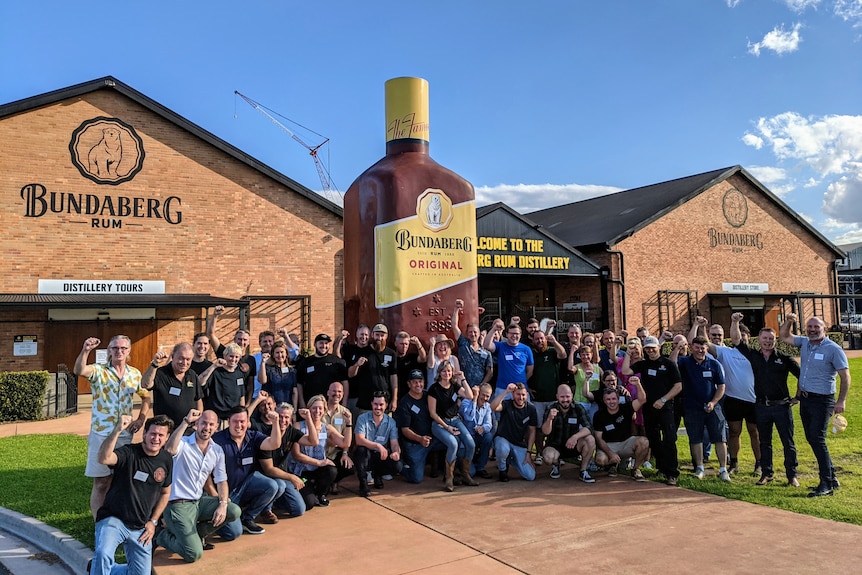 A large number of people pose for a photo outside the Bundaberg Rum distillery on a sunny day.