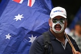 A man wears a mask with a hole cut out of it and carries the Australian flag