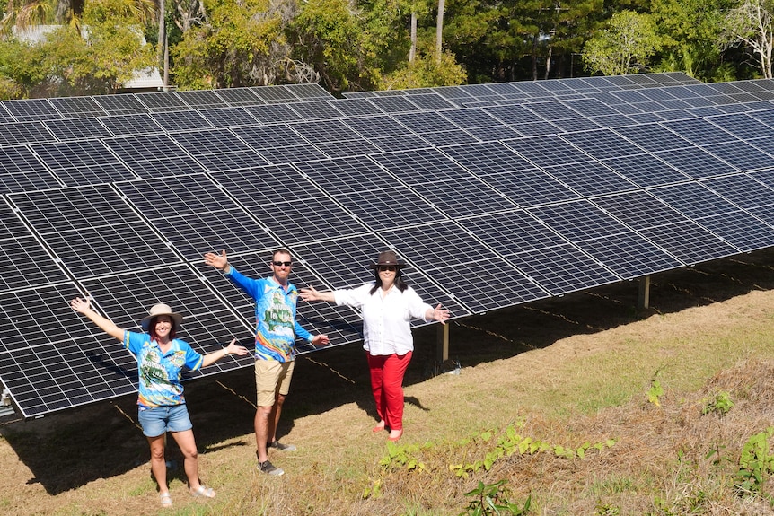 Three people stand in front of a solar farm with their arms stretched out.