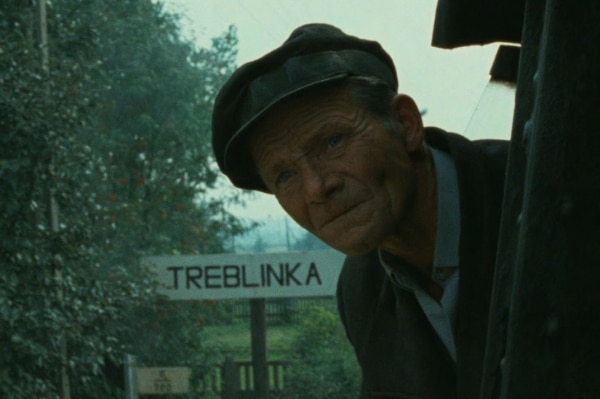A man leans out from a train with a sign reading 'Treblinka' in the background.
