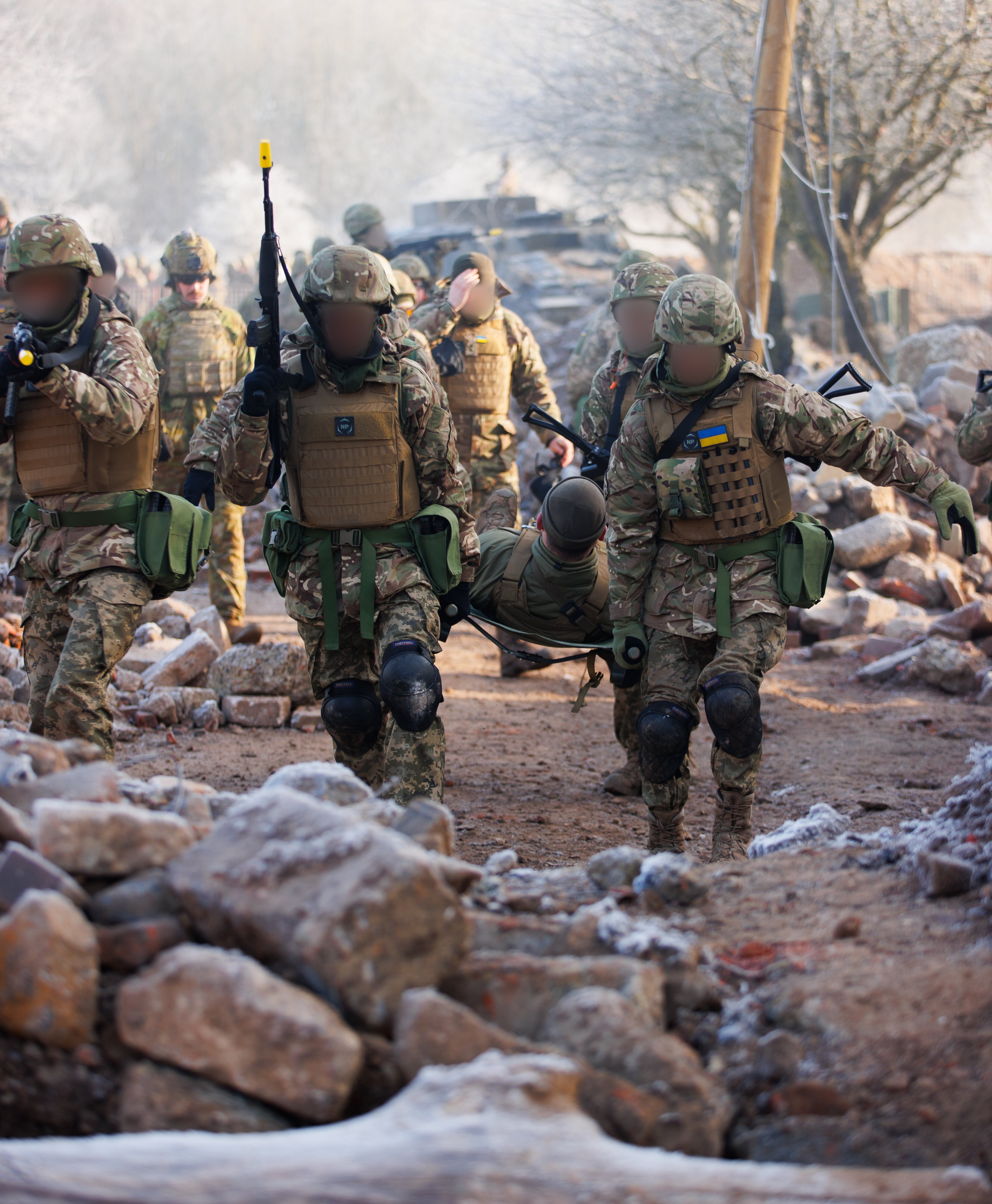 A group of soldiers carrying a person lying on a stretcher towards the camera, surrounded by rubble