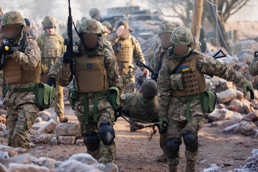 A group of soldiers carrying a person lying on a stretcher towards the camera, surrounded by rubble