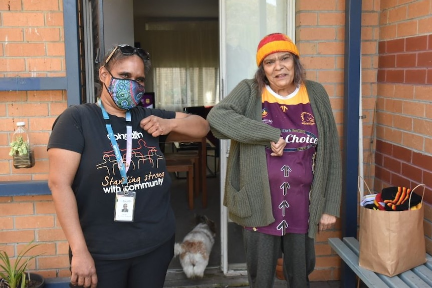 Two women stand on the front doorstep of a house and touch elbows, in a greeting synonymous with the coronavirus pandemic.