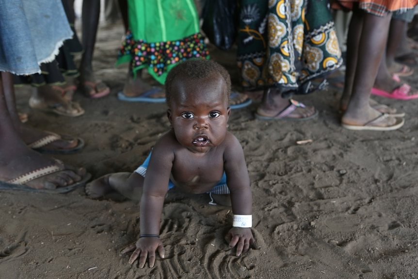 A toddler from South Sudan plays in the dirt at a refugee camp. People's feet are visible in the background.