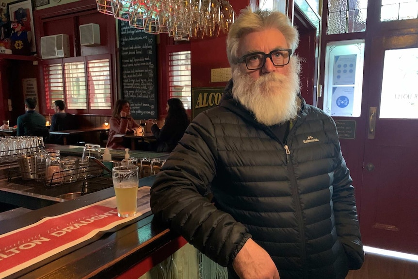 A man with a beard and glasses leans on a bar.