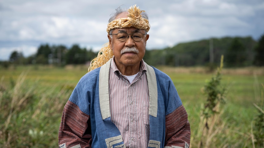 A man in a woven headdress stands in a field, looking solemn 