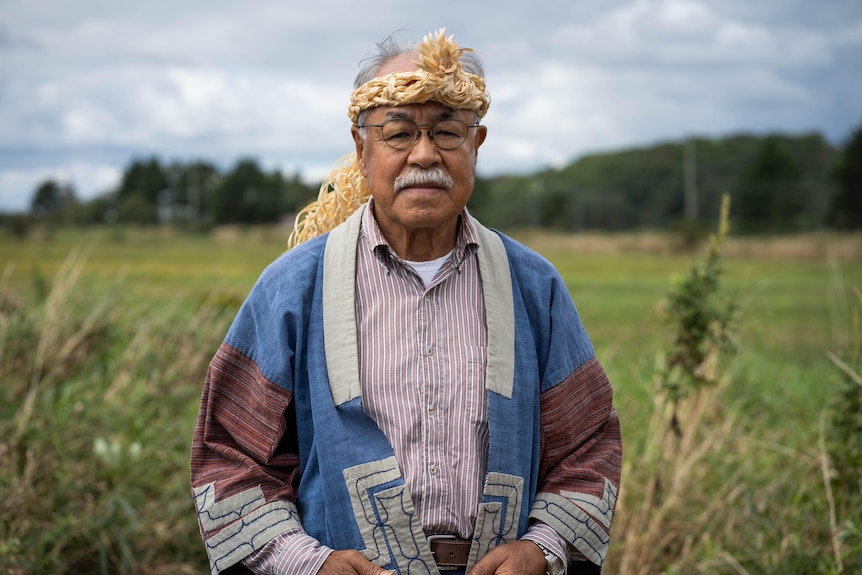 A man in a woven headdress stands in a field, looking solemn 