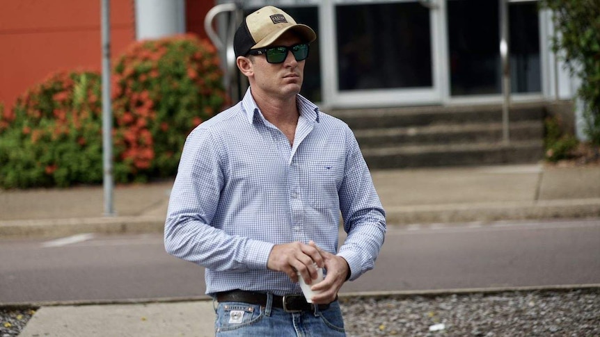 A man in a baseball cap, sunglasses and collared shirt, holding a coffee cup, crossing the road.