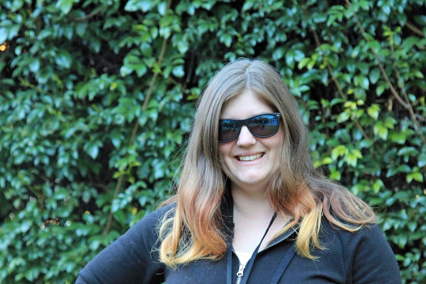 Woman wearing dark glasses standing in front of a hedge and smiling