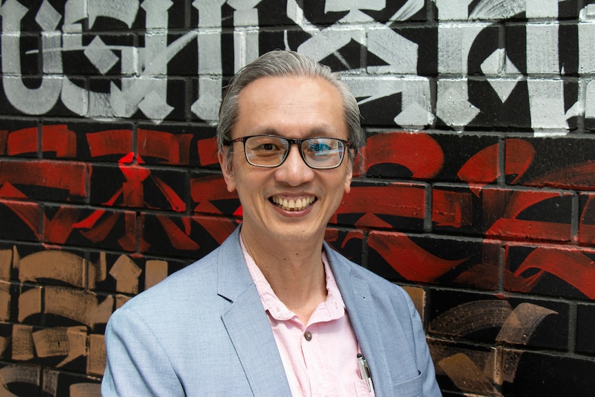 A man wearing a grey blue suit and pink business top, glasses smiling outside in front of street art 