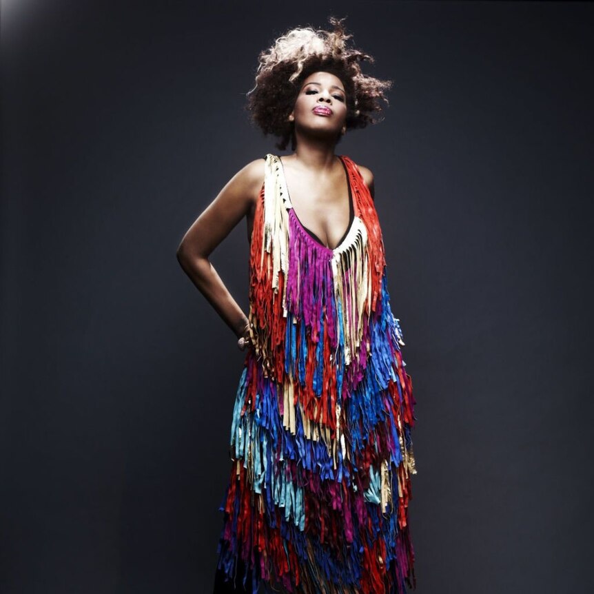 Macy Gray in a purple, gold and red tassled top, hand on hip, mouth closed, natural hair
