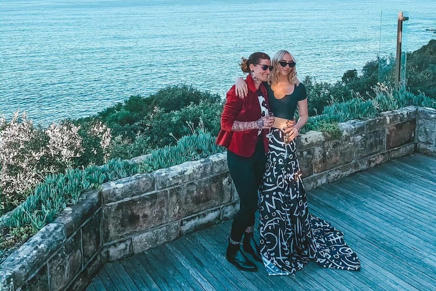 Two women stand holding champagne glasses on a balcony overlooking the ocean.
