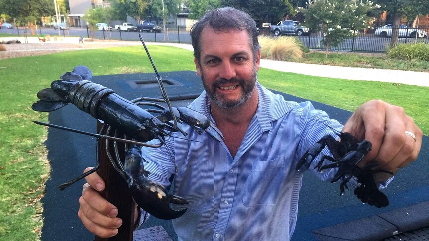 A man holds up a trophy with a fake yabby while holding a real yabby in his other hand.