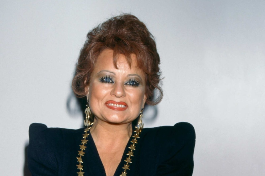 portrait of tammy faye grinning with glam makeup and updo, wearing black outfit and heavy gold jewellery