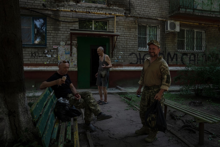 Three men laugh and eat outside a ruined and graffitied  building. Two are in military uniforms. 