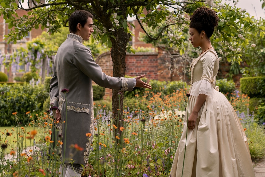 George holds out his hand for Charlotte to shake as they stand in a garden surrounded by tall flowers.