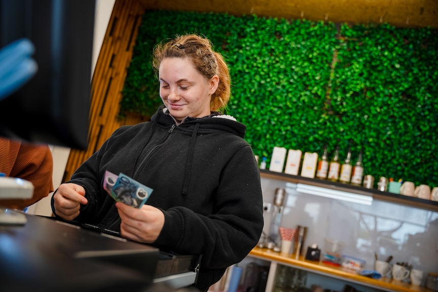 A young woman in a black jacket stands behind a cash register, sorting cash and smiling.