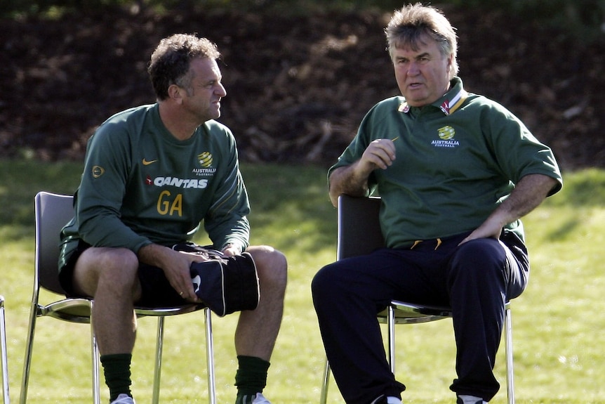 Two middle-aged men in coaching gear sit chatting on metal chairs, with green grass behind them and shadows from unseen trees