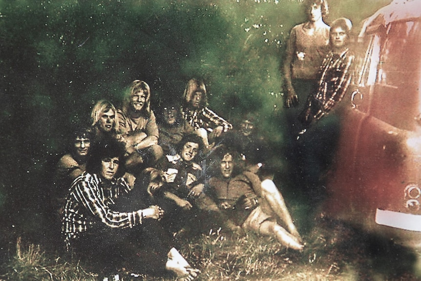 a 1970s photo group of young men with surfie hair reclining on the grass next to a van.