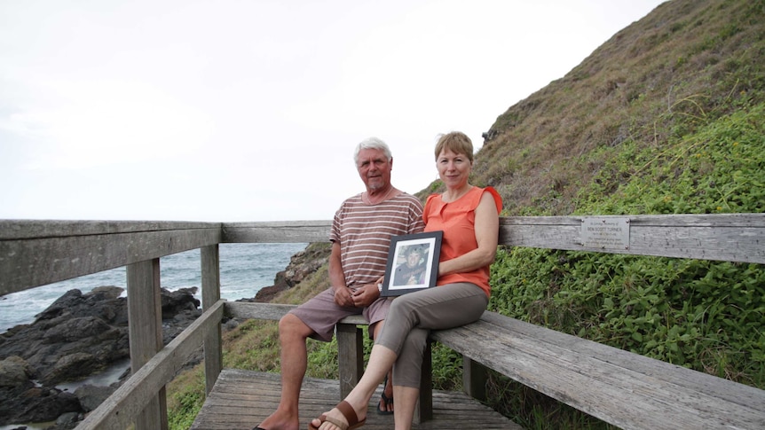 Lyn and Pete Turner sitting on an outdoors bench, holding a framed photograph of their son.