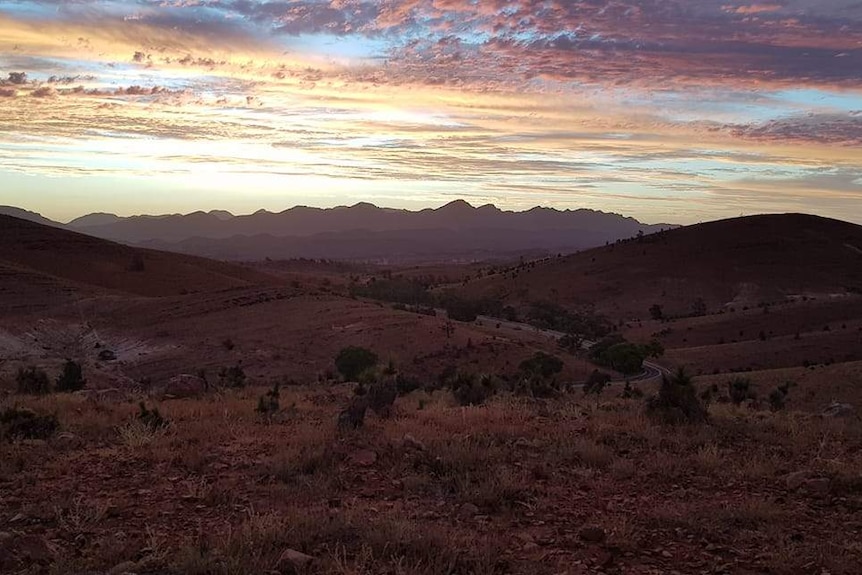 Bushland extends into the horizon where the sun sets behind large hills.