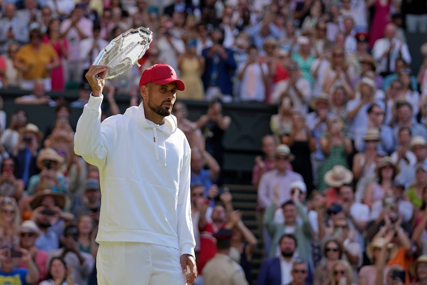 Nick Kyrgios is wearing a white hoodie and a red cap, holding up the runners-up trophy