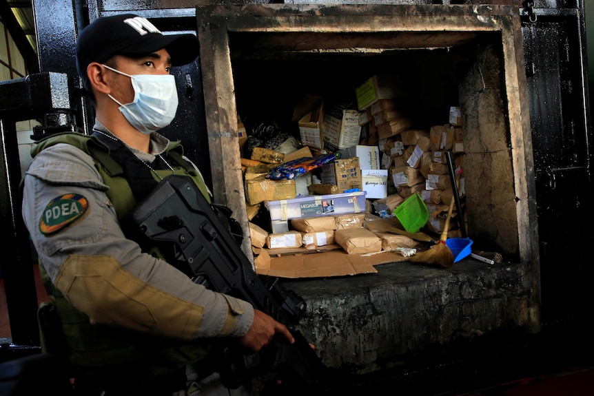 A man in camo gear holding a huge machine gun stands next to a hole in a wall