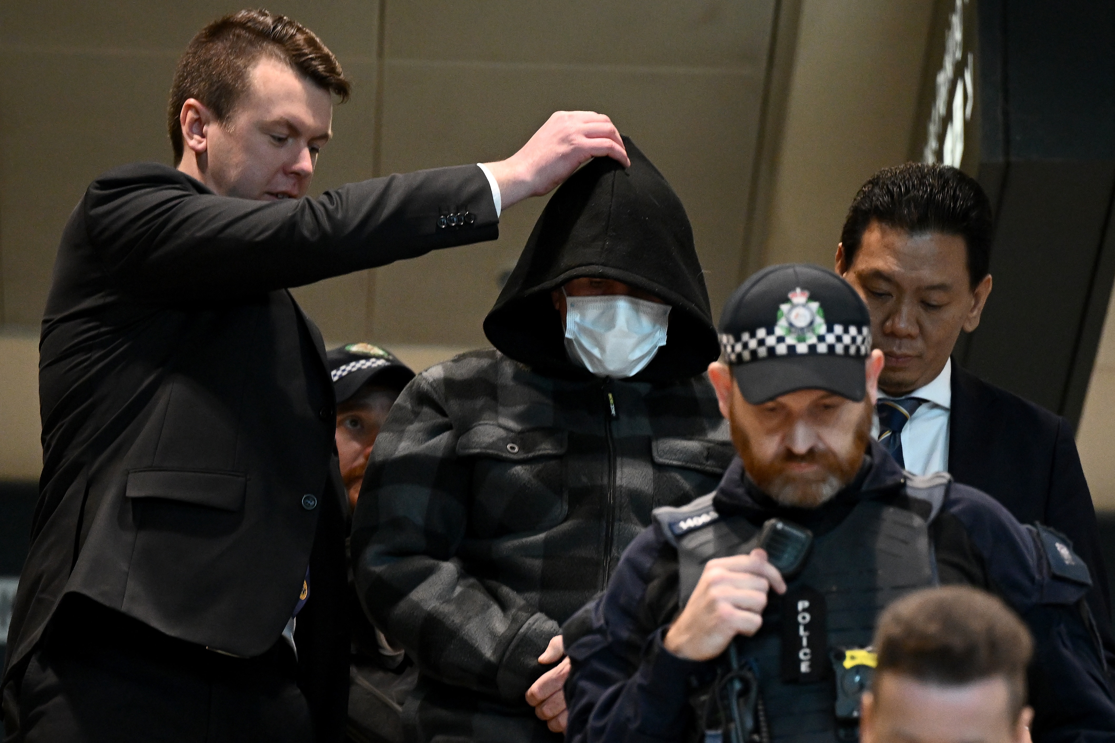 Man in black hood and jacket wearing face mask escorted by police.