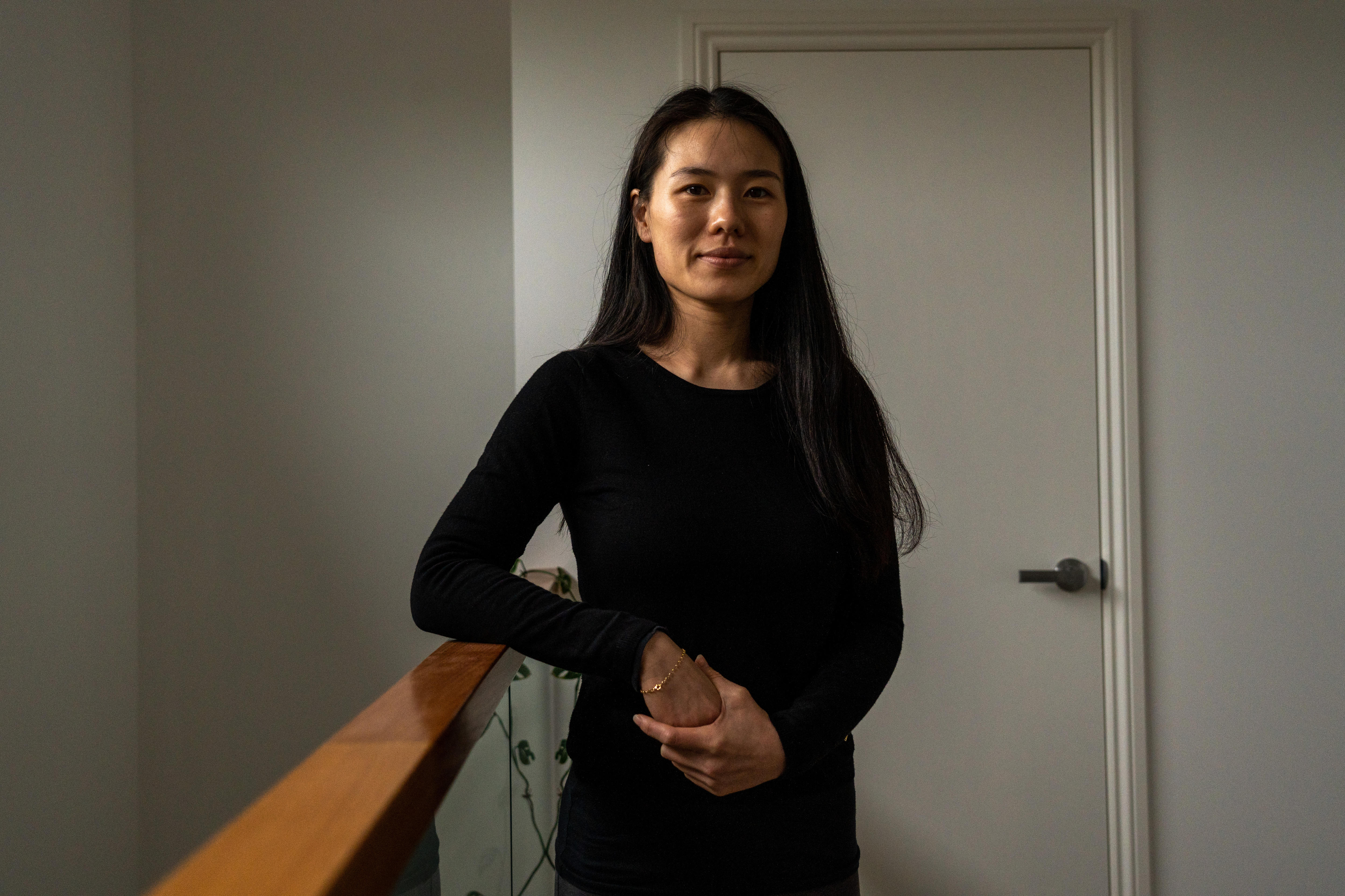 Joanne wears a black top and has long black hair and stands at the top of a staircase looking at the camera.