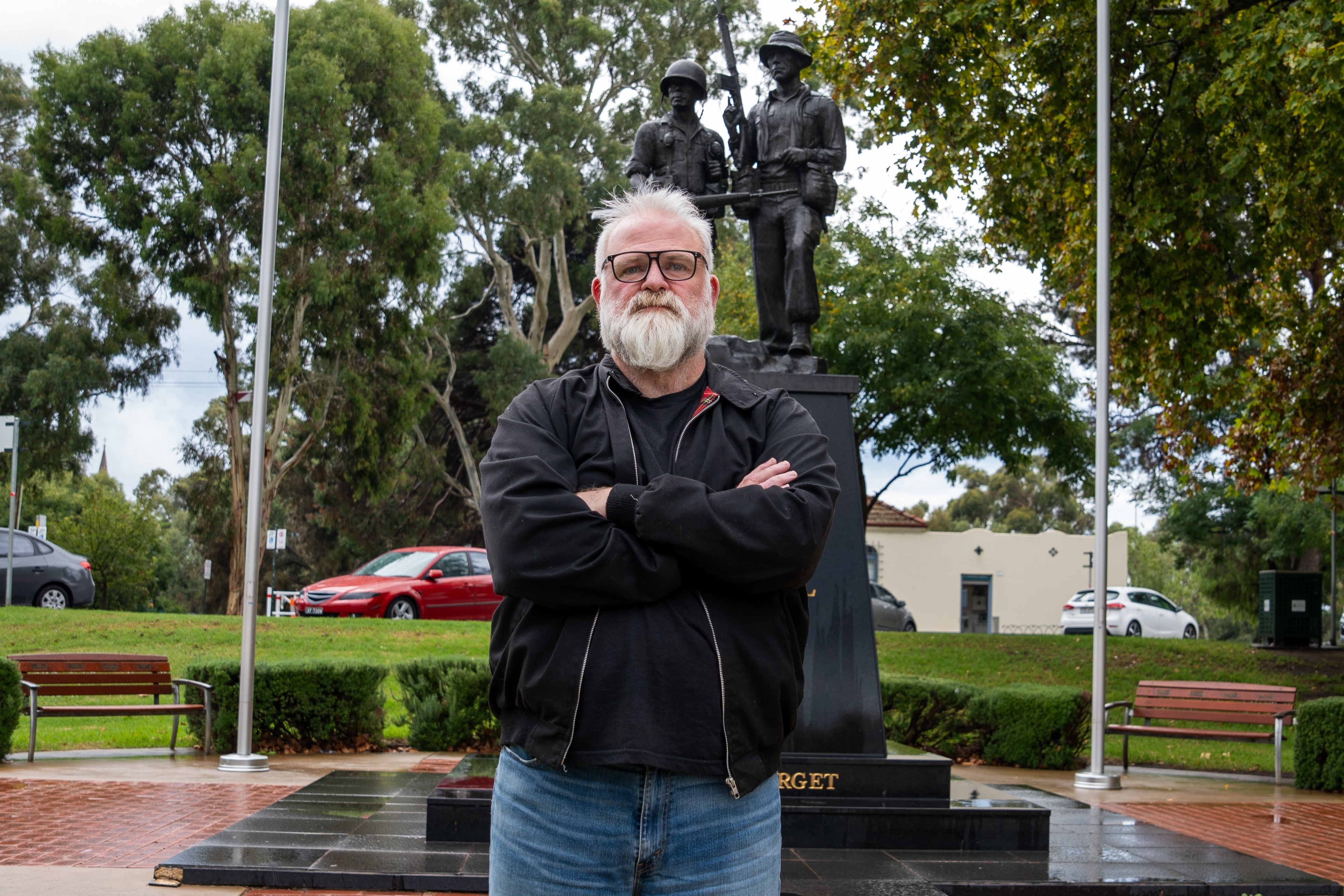 Un hombre de pelo blanco y barba se encuentra frente a un monumento a los caídos con los brazos cruzados.