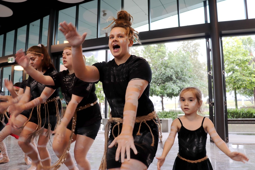 A line of Indigenous dancers dressed in black t-shirts and shorts perform in a hall