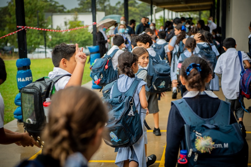 A crowd of school kids wearing pale blue uniforms and green backpacks walk to class, one girl looks back into the camera.
