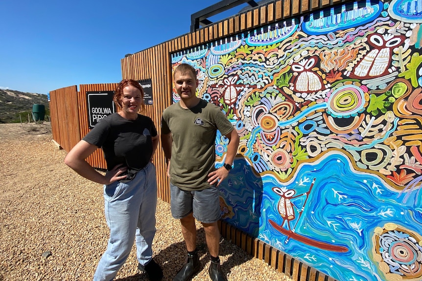 A woman and a man stand in front of a cafe with a mural on a wall