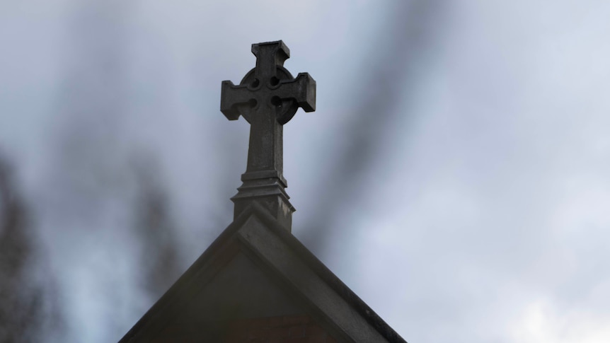 A close up of a stone cross on top of a church roof with grey background.
