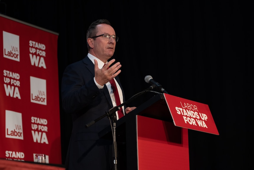 A wide shot of Mark McGowan wearing glasses standing at a lectern in front of WA Labor party signage.