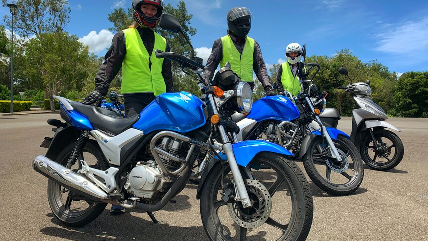 Three learner riders stands beside motorbikes at a training centre.