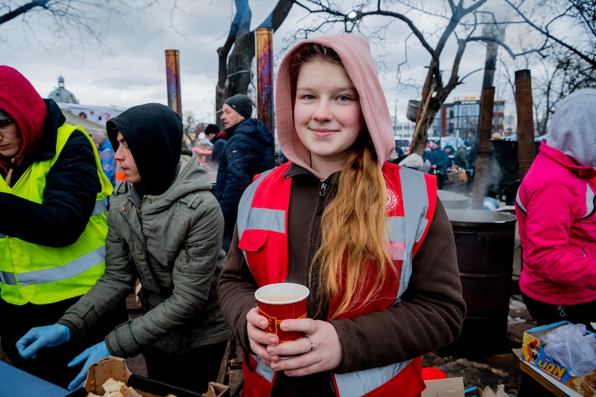 A teenage girl in winter clothes and a high-viz vest smiles while holding a paper cup of soup, standing near other volunteers.